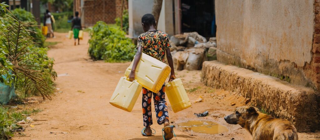 A child walks down a dirt path carrying two large yellow water containers, with more people and a dog visible along the path in a rural setting.