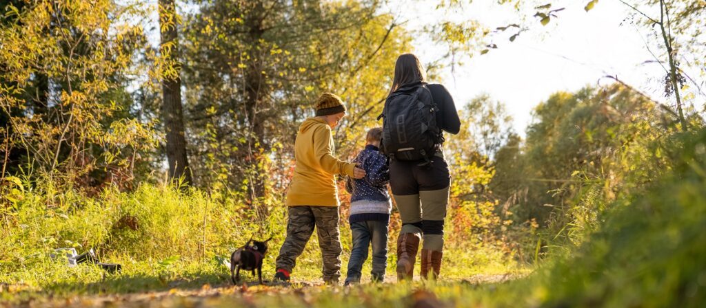 A woman with a backpack walks on a forest trail with two children and a small black dog on a sunny day. The group is surrounded by trees and autumn foliage.