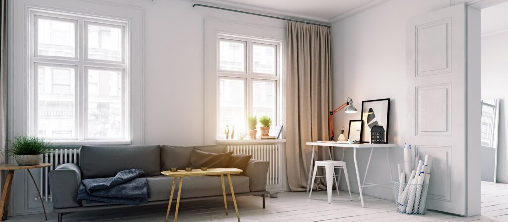 Bright living room with large windows, a gray sofa, side tables with plants, a cozy throw, and a modern white desk with a computer, lamp, framed art, and stationery. Neutral tones and natural light fill the space.