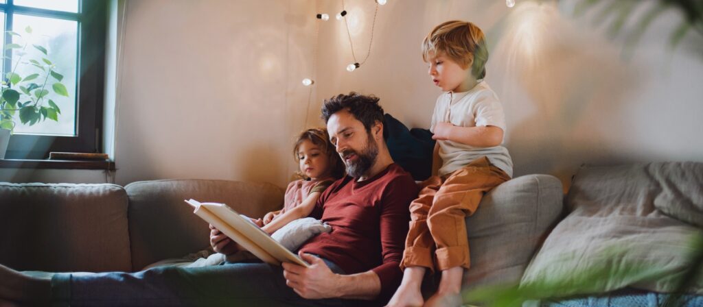 A man sits on a couch reading a book to two young children, one snuggled beside him and the other leaning over his shoulder. Warm lights and plants create a cozy, relaxed atmosphere in the living room.