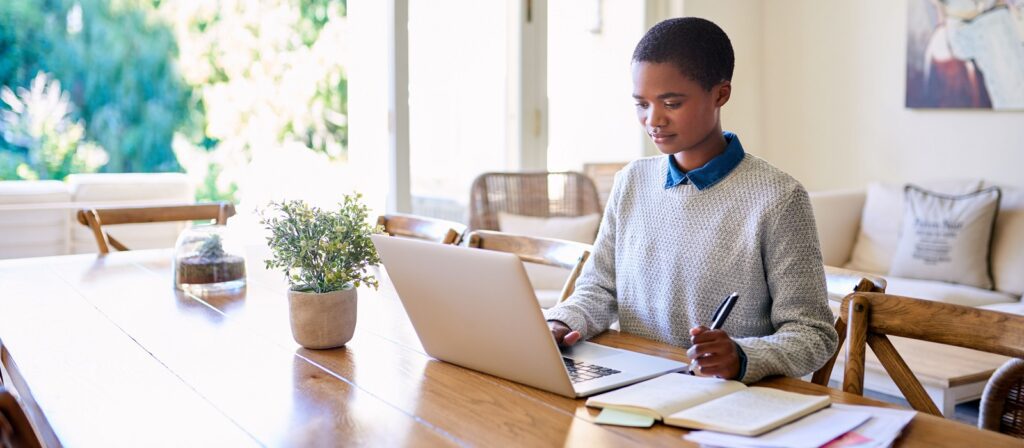 A person sits at a wooden table using a laptop, holding a pen and writing in a notebook. Sunlight streams in through large windows, and a potted plant and books are on the table. The setting appears calm and focused.