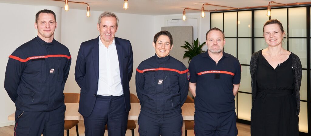 Five people stand indoors, three in dark blue uniforms with red stripes, and two in business attire. They are smiling, standing in a well-lit modern room with a wooden table and chairs behind them.