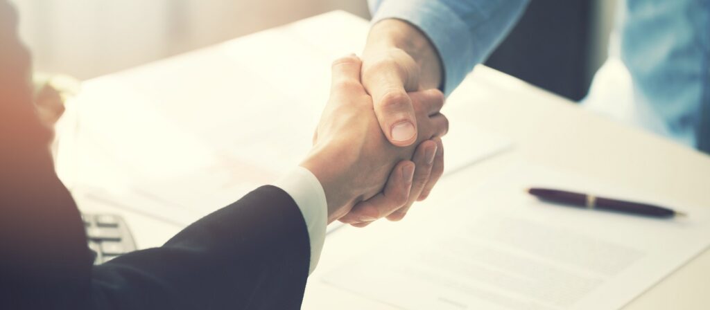 Two people shaking hands across a desk with documents and a pen, suggesting a business agreement or partnership.