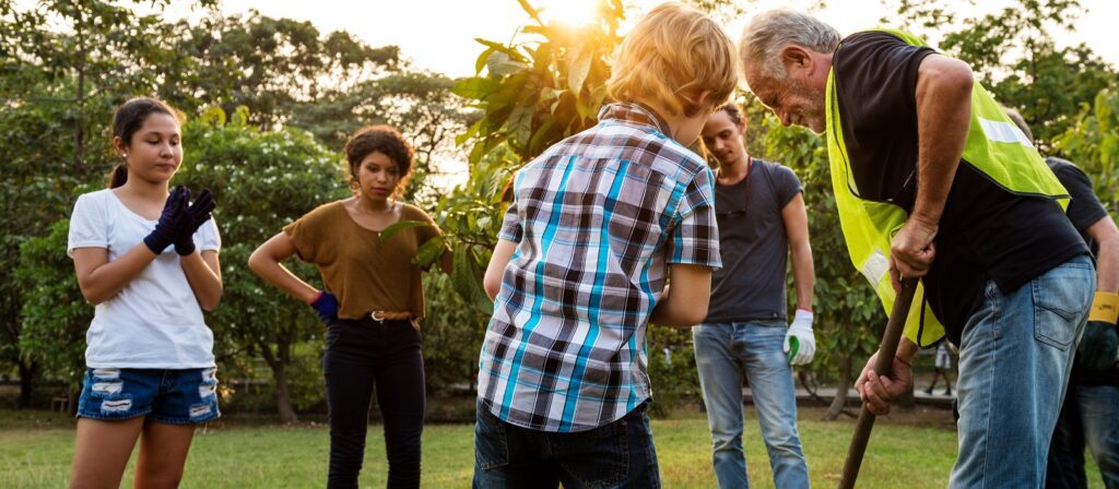 A group of people, including children and adults, work together outdoors planting a tree. One older man digs with a shovel while others watch and assist, surrounded by greenery in the sunlight.