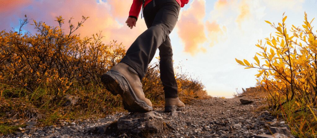 A person wearing hiking boots and dark pants walks up a rocky trail surrounded by autumn foliage, with a colorful sky in the background. The photo is taken from a low angle behind their feet.