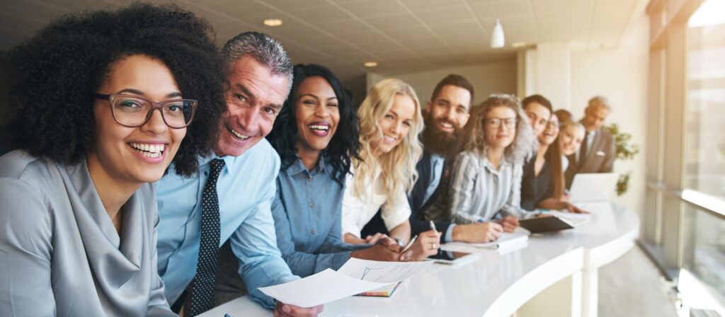 A diverse group of professionals, both men and women, are standing in a row at a modern office table, smiling and holding papers, with large windows in the background letting in natural light.