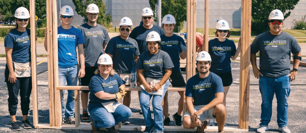 A group of volunteers wearing safety gear and Volunteer shirts pose together outdoors at a construction site, standing or kneeling in front of a wooden frame structure.