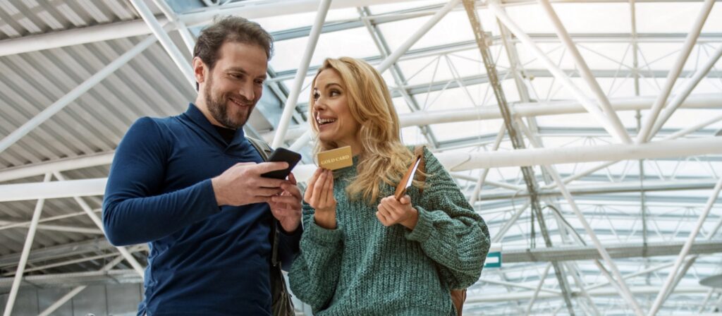 A smiling man and woman stand together in a modern airport terminal. The man looks at his phone while the woman holds up a gold card and a passport, both appearing happy and excited.