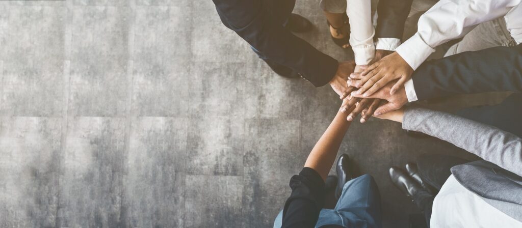 A group of diverse people dressed in business attire stand in a circle and stack their hands together in a gesture of teamwork and unity, viewed from above on a gray textured floor.