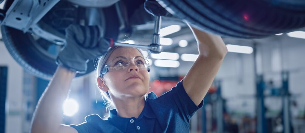 A mechanic wearing safety glasses and gloves uses a ratchet wrench to work on the underside of a car in a well-lit auto repair shop.