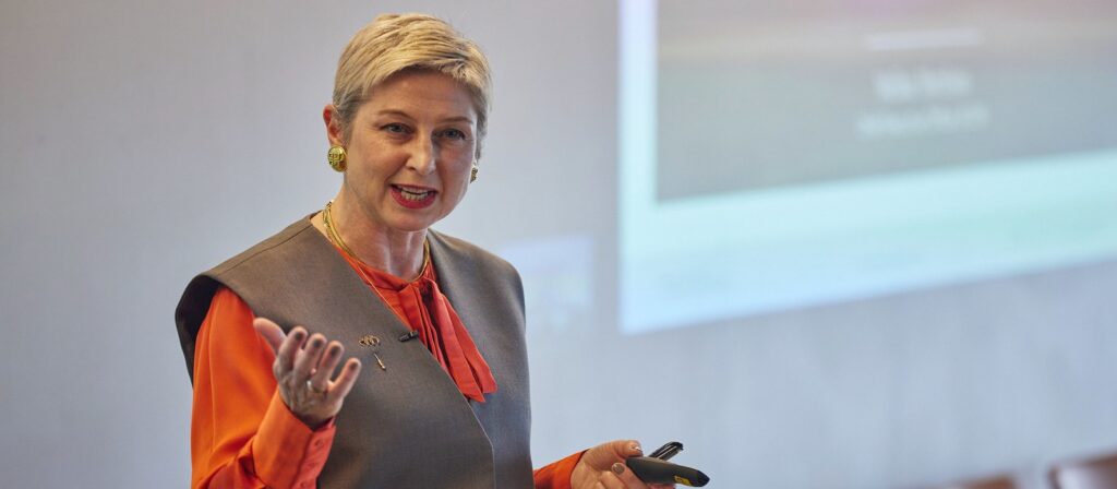 A woman with short blonde hair, wearing a grey vest over an orange blouse, stands and gestures while speaking in front of a blurred presentation screen.