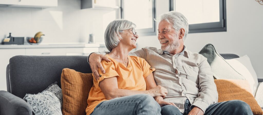 An older couple sits close together on a couch, smiling and looking at each other lovingly in a bright, modern living room.