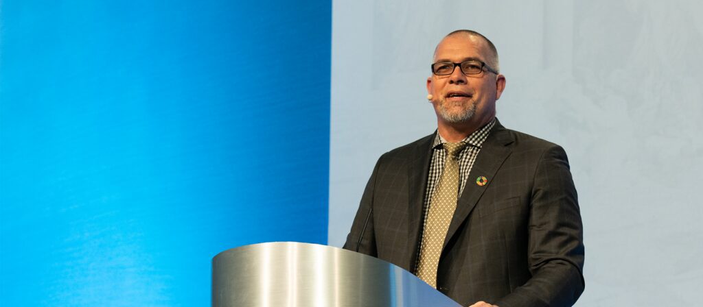 A man in a suit and tie stands at a podium, speaking into a headset microphone. The background is blue and white.