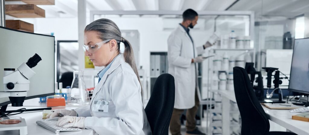 A woman scientist wearing safety goggles and gloves works at a lab desk near a microscope, while a man in a lab coat works in the background surrounded by laboratory equipment and shelves.