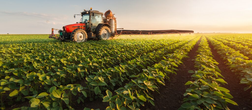 A red tractor sprays crops in a large, lush green field under a bright, clear sky at sunset, illustrating modern farming and agricultural technology.