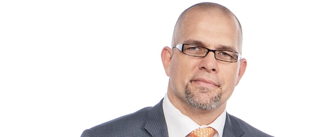 A man with a shaved head, glasses, and a short goatee wears a gray suit, white shirt, and orange patterned tie, standing against a plain white background.