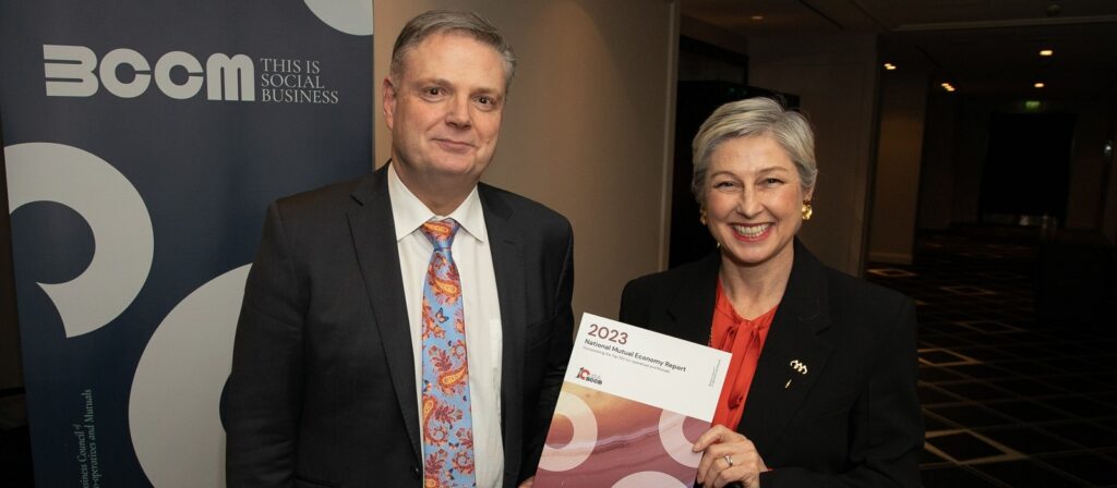 Two people in formal attire stand indoors. The woman on the right smiles and holds a 2023 award certificate, while the man on the left stands next to a branded banner that reads “SOCIAL BUSINESS”.
