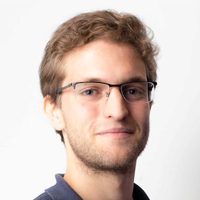 A young man with short, light brown hair and a trimmed beard is wearing rectangular glasses and a dark collared shirt, smiling slightly against a plain white background.