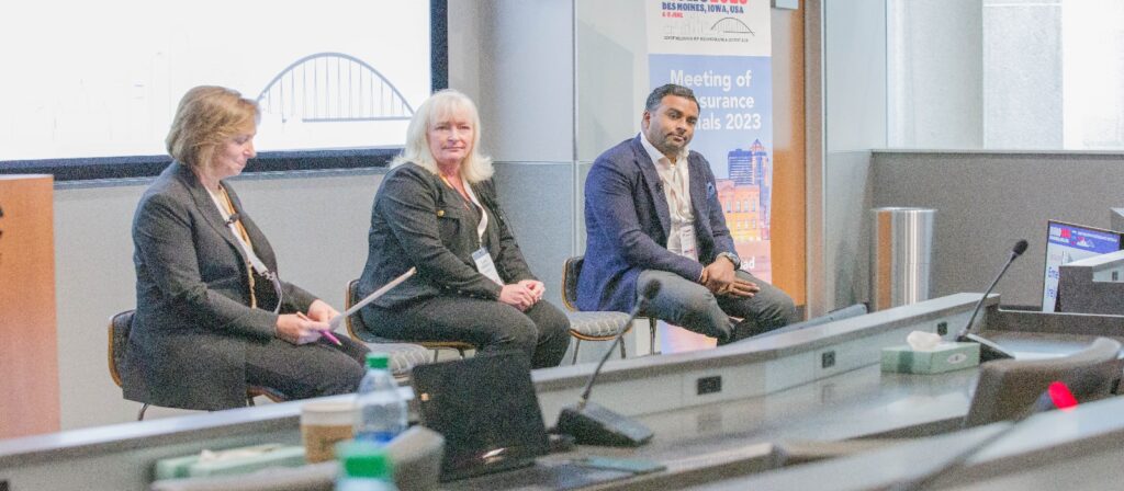 Three panelists, two women and one man, sit on stools at the front of a conference room during the Meeting of Insurance Minds 2023 event, speaking to an audience. A sign and screen are visible in the background.