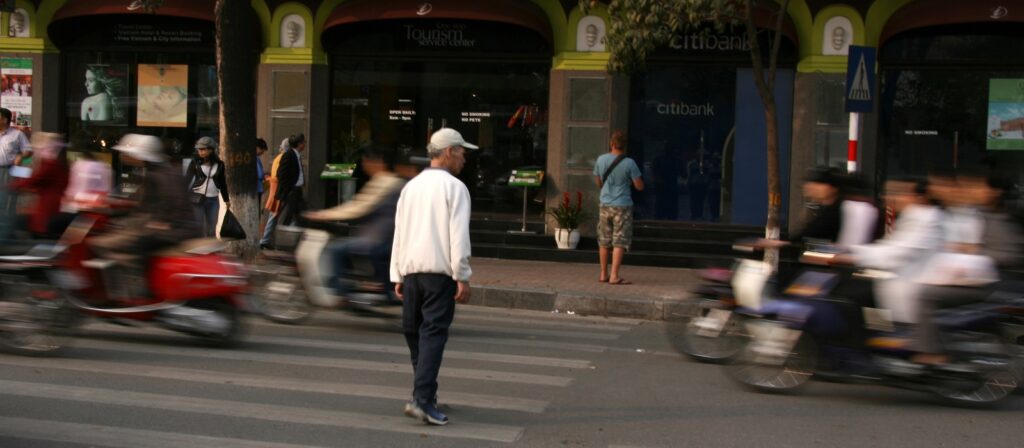 An older man walks slowly across a busy street while blurred motorbikes pass by, illustrating motion and urban life; a person stands at ATMs on the sidewalk in the background.