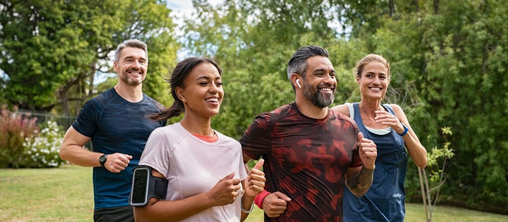 Four adults, two men and two women, are jogging together outdoors in a park. They are smiling, wearing athletic clothing, and look happy and energetic. Trees and greenery are visible in the background.