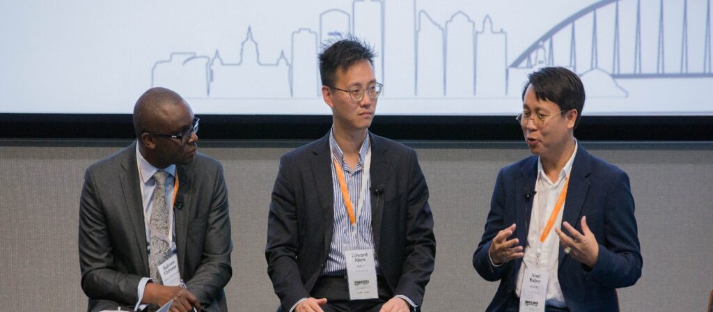 Three men in business attire sit and talk on a panel. Two listen attentively while the third gestures as he speaks. They wear conference name badges and orange lanyards. A cityscape graphic is visible in the background.