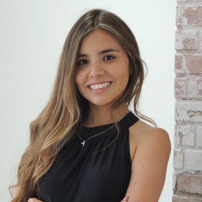 A young woman with long brown hair, wearing a sleeveless black top and a necklace, smiles while standing in front of a white wall with exposed brick.