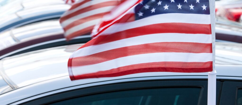 Close-up of an American flag on a car, with several other flags and cars visible in the background, suggesting a patriotic event or car dealership promotion.
