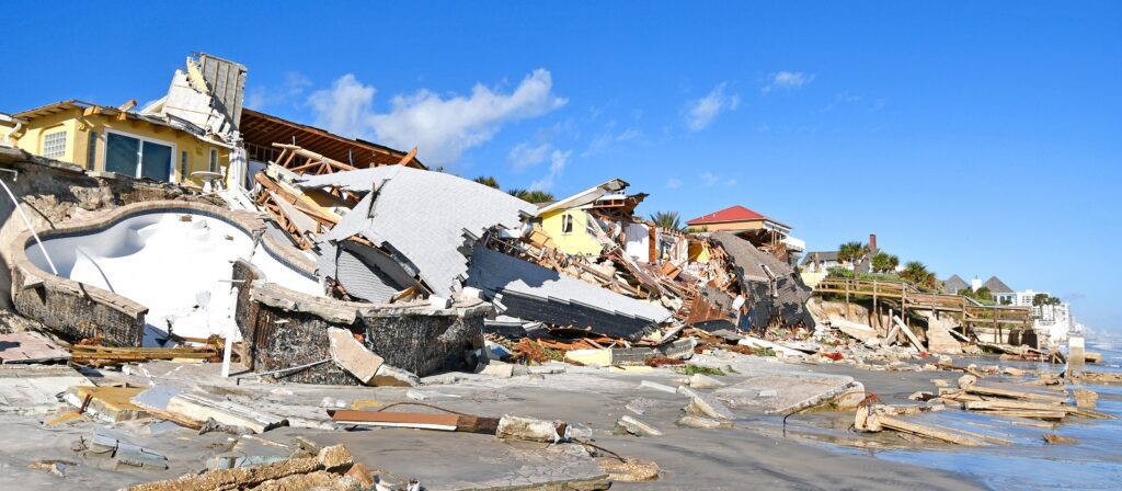 Damaged beachfront homes and debris scattered along the shore after a natural disaster, with collapsed roofs, broken structures, and a destroyed swimming pool under a clear blue sky.