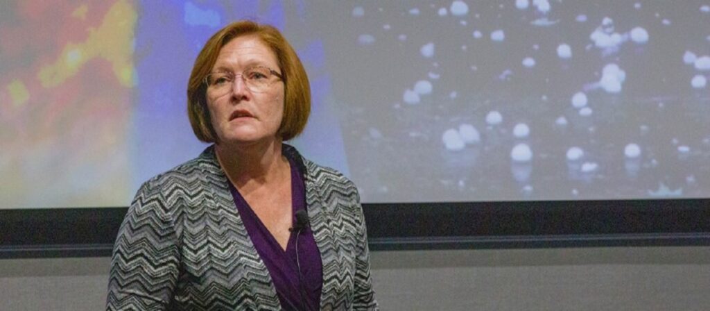 A woman with short reddish hair and glasses stands in front of a large screen displaying colorful abstract shapes and white spots, wearing a chevron-patterned blazer over a purple top.