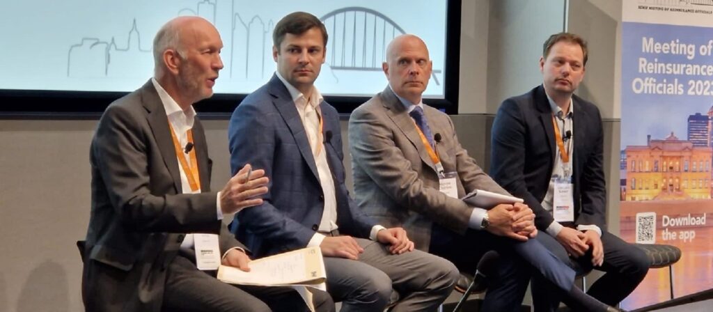 Four men in business attire sit on a panel at a conference, speaking to an audience. Behind them are a screen with a cityscape graphic and a banner that says Meeting of Reinsurance Officials 2023.