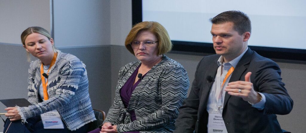 Three people sit at a panel discussion. A man on the right gestures while speaking, while two women listen attentively. All are wearing conference name badges and business attire. A projection screen is visible behind them.