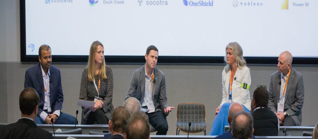 Five people sit on a panel discussion in front of an audience, with a screen behind them displaying company logos. Four men and one woman are engaged in conversation, each wearing business attire and conference lanyards.