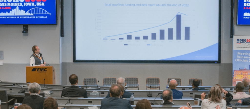 A presenter stands at a podium in a conference room, showing a slide with a bar and line graph to an audience seated at desks. The slide discusses insurtech funding and deal count up to the end of 2022.