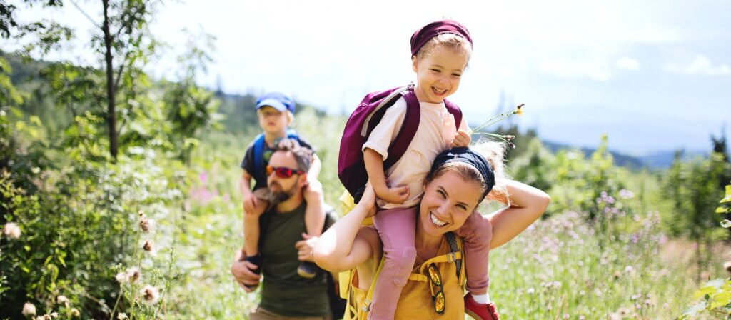 A smiling woman gives a young girl a piggyback ride while a man carries a young boy on his shoulders. They are hiking outdoors on a sunny day, surrounded by greenery and wildflowers.