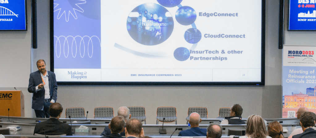 A speaker presents to an audience in a lecture hall with a slide displaying business topics like EdgeConnect, CloudConnect, and InsurTech partnerships at an EMC Insurance Companies 2023 event.