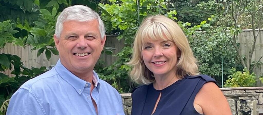 A man and woman smiling outdoors in a garden with green plants and a wooden fence in the background. The man wears a light blue shirt, and the woman wears a dark sleeveless top.