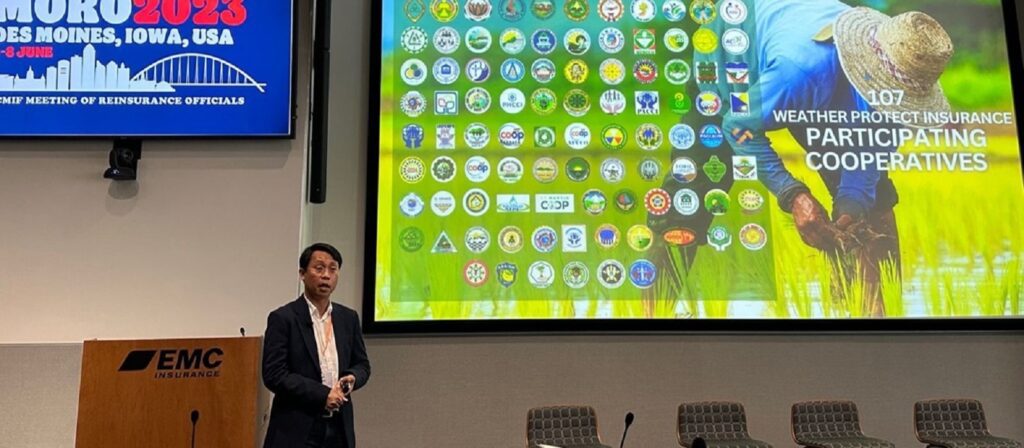 A man in a suit stands presenting in front of a large screen showing agricultural logos and a farmer planting rice. A conference banner reads IMURO 2023, Des Moines, Iowa, USA. Four empty chairs are visible near the stage.