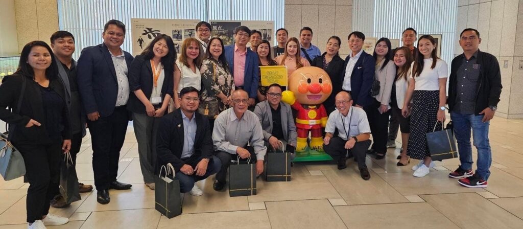 A group of people pose and smile together indoors, standing and crouching around a large statue of the cartoon character Anpanman, with some holding gift bags.