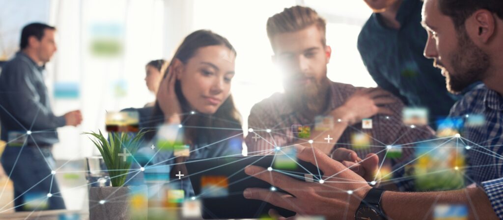 A group of young adults gathers around a tablet in a modern office, with digital network graphics overlaying the image, symbolizing technology and collaboration. Others converse in the background.