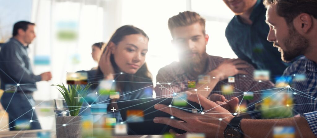 A group of young adults collaborate around a tablet at a desk, with digital network graphics and icons overlaid, suggesting technology and teamwork in a modern office setting.