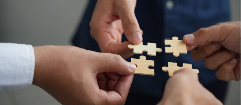 Four hands, each holding a wooden puzzle piece, are coming together to connect the pieces, symbolizing teamwork and collaboration.