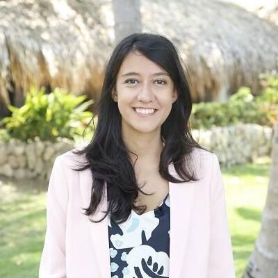 A woman with long dark hair, wearing a light pink blazer and a patterned top, stands outdoors in a sunny, lush garden with thatched-roof structures and greenery in the background. She is smiling at the camera.