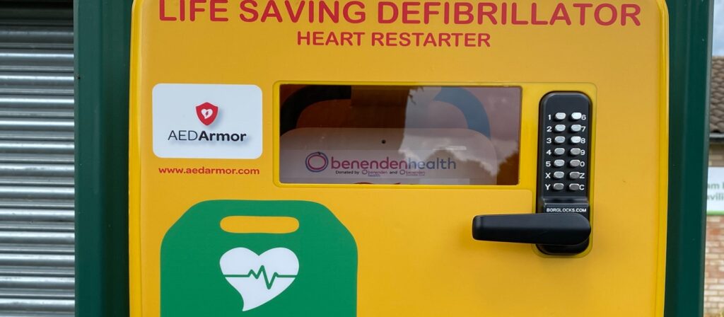 A yellow life-saving defibrillator unit with a keypad and window, labeled “LIFE SAVING DEFIBRILLATOR HEART RESTARTER” and featuring AED and heart icons. Logos for AEDArmor and Benenden Health are also visible.