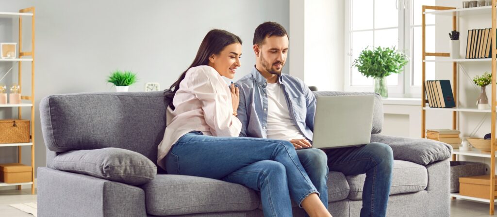 A young couple sits together on a gray couch in a bright living room, smiling and looking at a laptop on their laps. Shelves with books, plants, and decor are visible in the background.