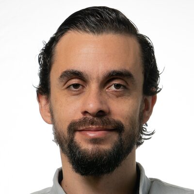 A man with dark hair slicked back, a beard, and mustache, wearing a light-colored collared shirt, is looking at the camera with a neutral expression against a plain white background.