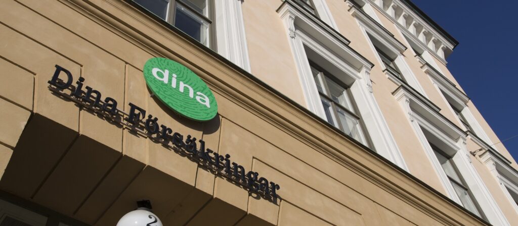 A beige building with white-trimmed windows and a green circular sign reading dina above the black text Dina Försäkringar on the facade, viewed from a low angle against a clear blue sky.