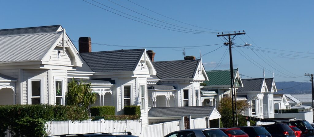 A row of white wooden houses with gabled roofs and chimneys lines a sunny street, with parked cars in the foreground and power lines overhead against a clear blue sky.