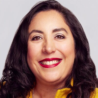 A woman with long dark hair smiles brightly. She is wearing red lipstick, subtle eye makeup, and a yellow top. The background is plain and light-colored.