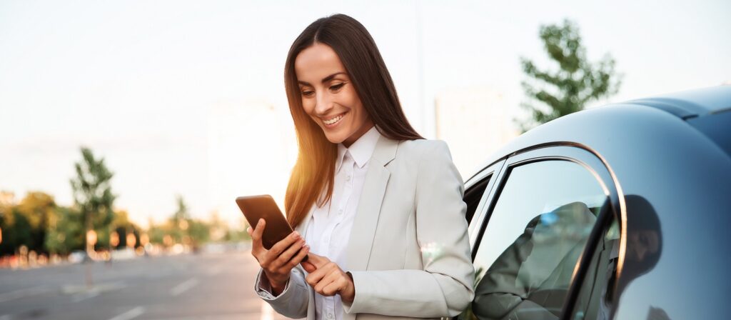 A woman in a white blazer stands by a car in a parking lot, smiling as she looks at her smartphone. Trees and blurred buildings are visible in the background, with sunlight shining behind her.